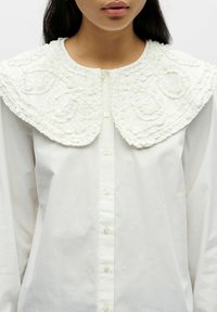 Camisa blanca de algodón con un gran cuello rizado texturizado y bordado detallado. Los botones delanteros son pequeños y discretos.