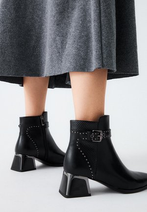 Botas de tobillo de cuero negro con un panel texturizado, acentos de tachuelas y un tacón metálico cuadrado. Combinadas con una falda gris.