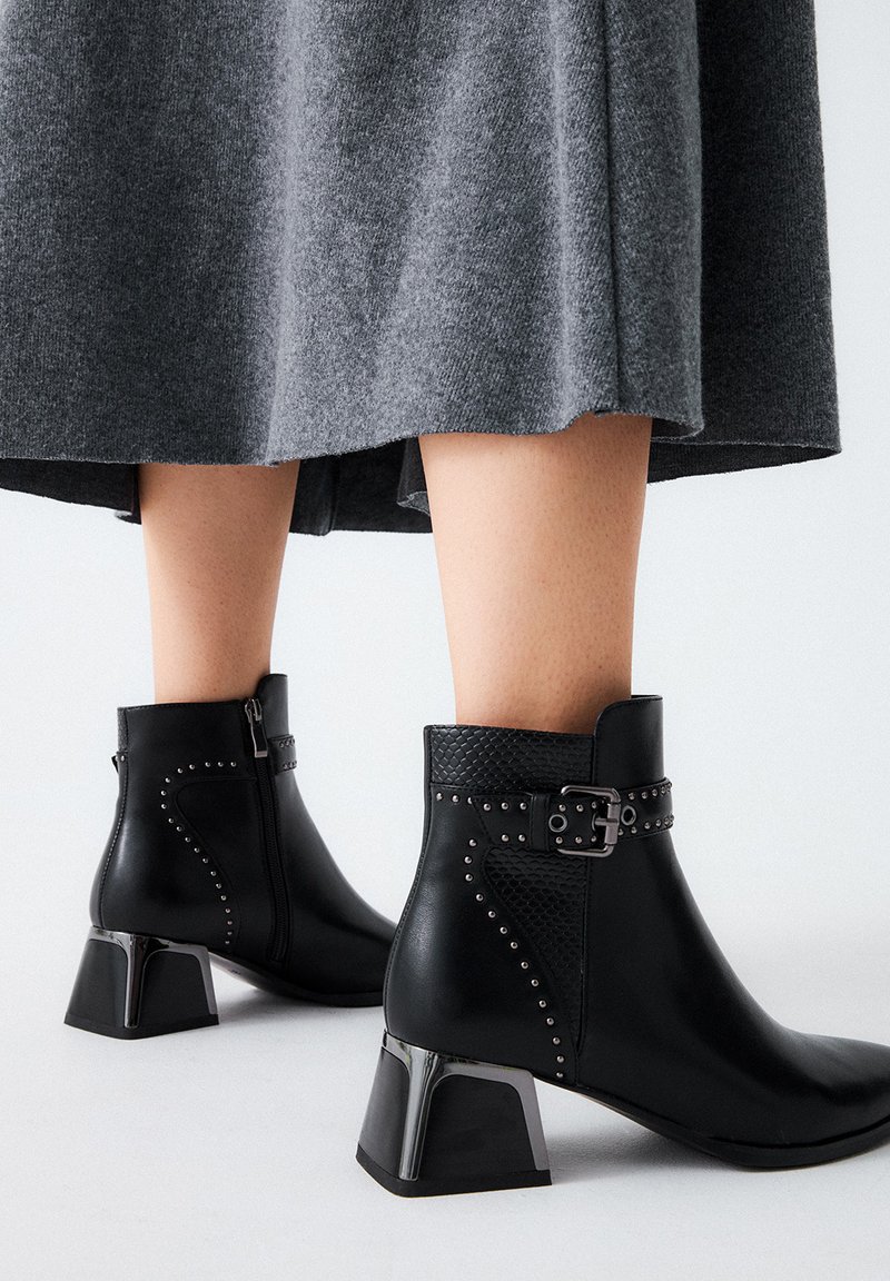 Botas de tobillo de cuero negro con un panel texturizado, acentos de tachuelas y un tacón metálico cuadrado. Combinadas con una falda gris.