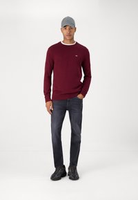 Pull en maille bordeaux avec un col rond, casquette grise, jeans foncés et chaussures noires. Tissu texturé et détail de col contrastant.