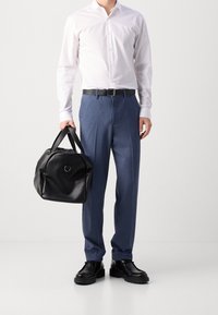 Witte overhemd met knopen, blauwe op maat gemaakte broek, zwarte leren riem en zwarte gestructureerde handtas. Model draagt zwarte nette schoenen.