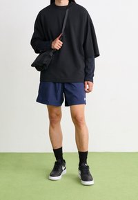 Person som bär en svart oversized sweatshirt, marinblå träningsshorts, svarta höga strumpor, svarta Nike-sneakers och bär en svart axelväska.