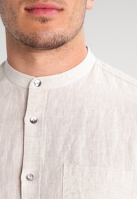 Camisa de lino en un beige claro con cuello mao. Presenta botones plateados y un bolsillo en el pecho izquierdo. Textura suave y diseño casual.