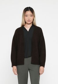 Mørkebrun cardigan med strukturstrikk, åpen front og avslappet passform. Brukt over en svart topp og parret med grå bukser.