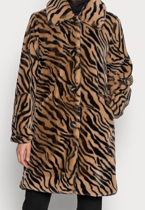Manteau en fourrure marron et noir à rayures de tigre avec grand col et boutons noirs, porté sur des vêtements noirs, main dans la poche droite.