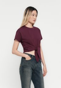 Bordeaux katoen t-shirt met korte mouwen, geknoopt in de taille. Gecombineerd met donkere spijkerbroek. Eenvoudig, aansluitend ontwerp met een gladde textuur.