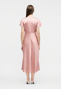 Γυναίκα που στέκεται με την πλάτη, φοράει μεταξωτό ροζ midi φόρεμα με κοντά μανίκια και λευκά mule τακούνια σε απλό φόντο.