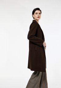 Ruskea villakangastakki, jossa on pitkä, suora siluetti, leveät läpät ja sivutaskut. Mallilla on päällään ruudulliset leveät housut. Yksinkertainen tausta.