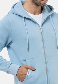Veste à capuche zippée bleu clair en tissu doux, dotée d'une poche avant, d'une capuche avec cordons ajustables et de poignets côtelés pour plus de confort.