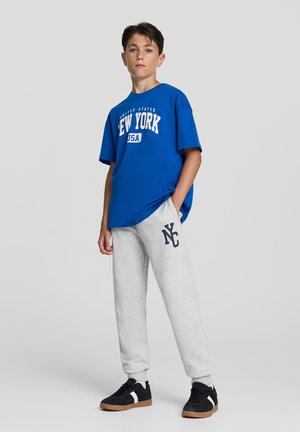T-shirt in cotone blu con stampa "New York", abbinata a pantaloni della tuta grigi con logo "NC". Sneakers nere con suole color sabbia.