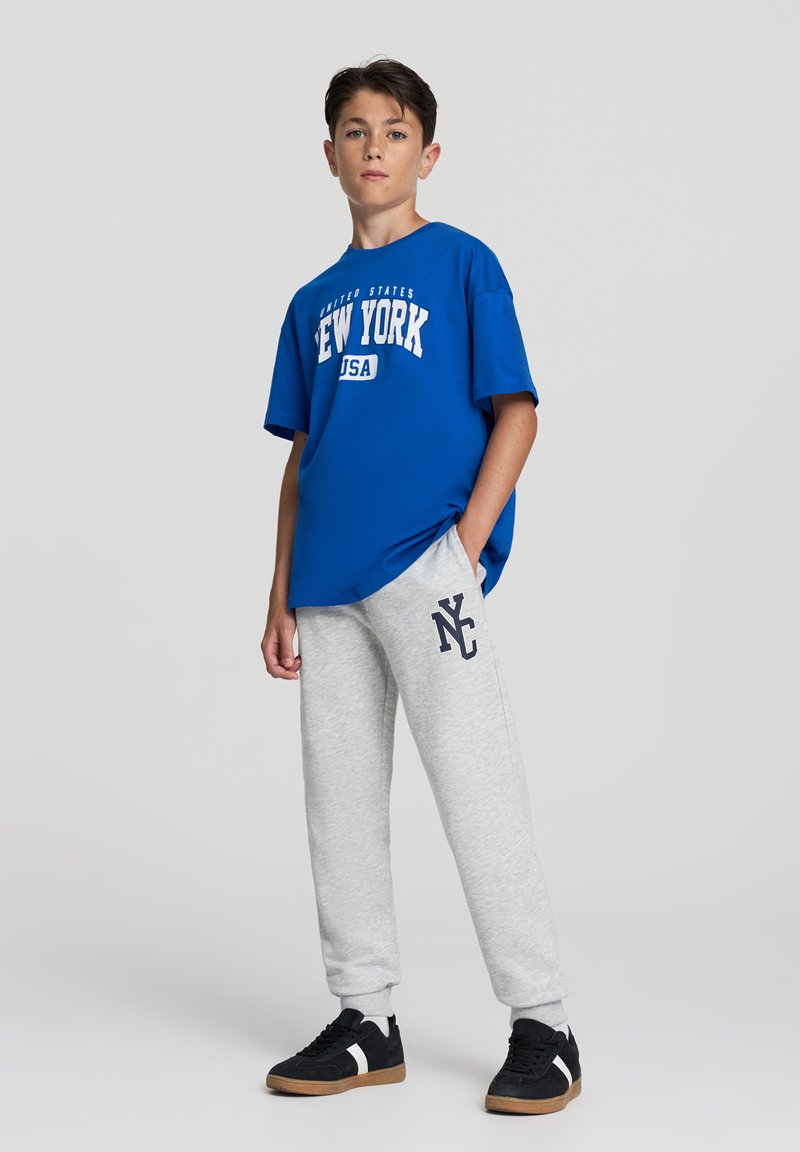 Niebieska bawełniana koszulka z nadrukiem "Nowy Jork", w połączeniu z szarymi dresami z logo "NC". Czarne trampki z beżowymi podeszwami.