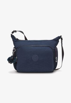 Sininen kangaskassi olkahihnalla, jossa on etupuolella vetoketjullinen tasku, teksturoitu pinta ja pyöreä logo. Kiinnitettynä on pehmeä apinamaski.