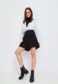 Schwarze hochgeschnittene Shorts mit Falten, kombiniert mit einem weißen Hemd mit Knöpfen und einem schwarzen Rollkragenpullover, ergänzt durch klobige schwarze Stiefeletten.