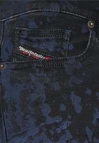 Dark denim jeans featuring a splatter pattern in shades of blue and black, with a visible back pocket and red and white brand label.