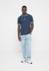 Marineblå polo med lynlås ved kraven, lyseblå detaljer på ærmerne. Parret med lyseblå denimjeans og hvide sneakers.