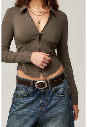 Kobieta w oliwkowej zielonej dopasowanej bluzce z długimi rękawami zapinanej na zamek, z nałożonymi na siebie złotymi naszyjnikami, pierścionkami, niebieskich dżinsach i brązowym pasku z owalną klamrą.