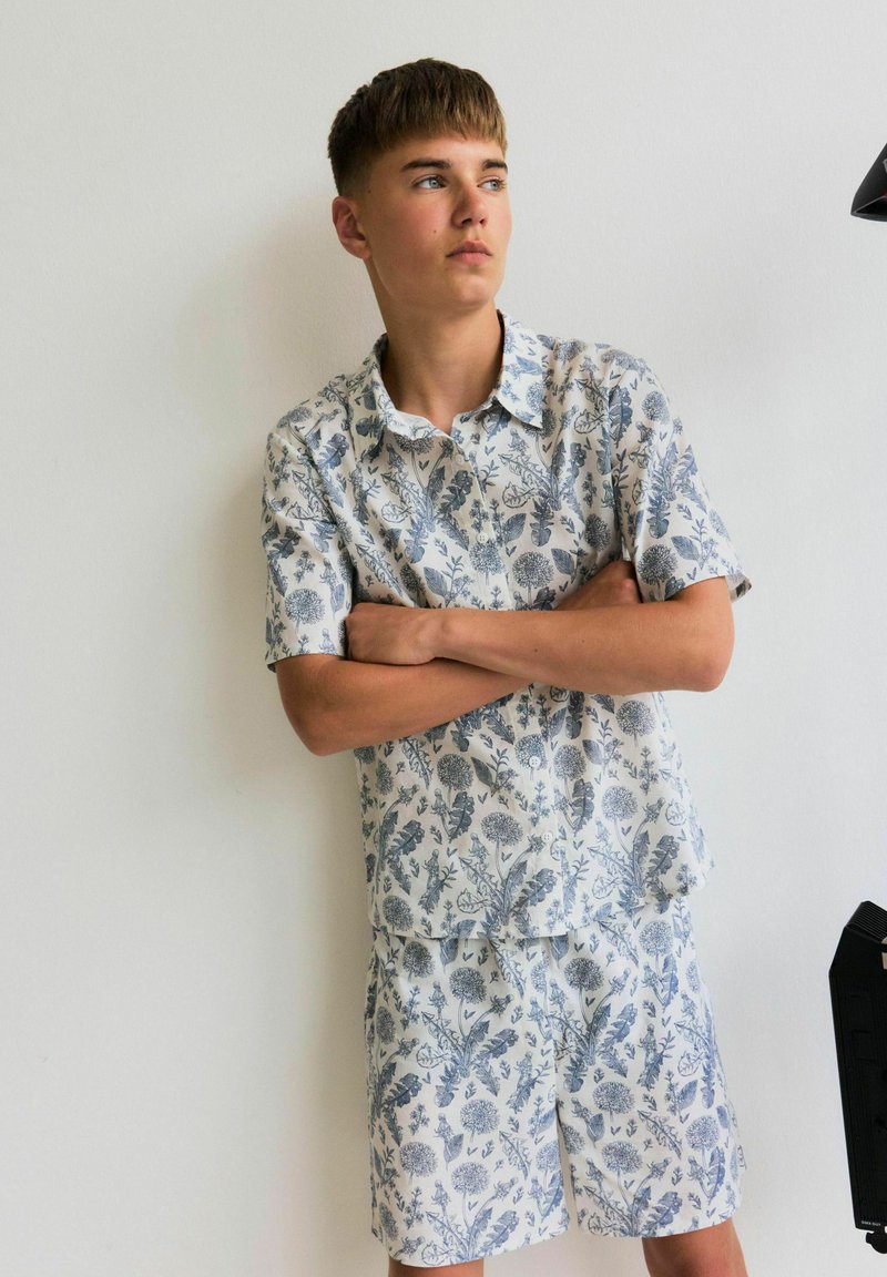 Ensemble de chemise à manches courtes et de short, avec un fond blanc et des motifs floraux bleus, fabriqué en tissu léger. Design décontracté.