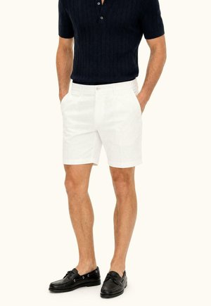 Uomo che indossa una polo blu navy a maniche corte, pantaloncini bianchi e mocassini neri, in piedi con le mani in tasca su uno sfondo chiaro e semplice.