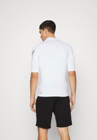 White fitted short-sleeve surf shirt with seamless design, worn with black shorts. Features a logo on the left shoulder. Smooth texture.