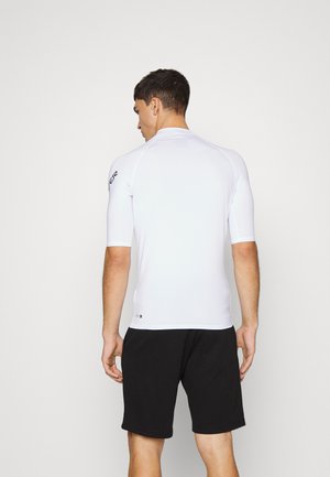White fitted short-sleeve surf shirt with seamless design, worn with black shorts. Features a logo on the left shoulder. Smooth texture.