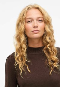 Brązowy dziany sweter z wysokim kołnierzem, z długimi rękawami i lekko bufkami na ramionach, noszony przez osobę o falujących blond włosach.