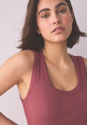 Jeune femme aux cheveux courts et foncés, portant un haut mauve sans manches côtelé, posant devant un fond pâle avec une expression neutre.