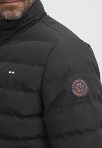Svart dunjakke med horisontale sømmer, høy krage og et rundt logo-patch på overarmen. Har en fargeblokkdetalj i rødt, hvitt og blått.