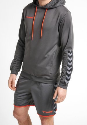 Felpa sportiva grigia con cappuccio e coulisse arancioni, tessuto texturizzato e accenti neri a forma di chevron sulle maniche. Abbinata a pantaloni corti grigi con finiture arancioni.