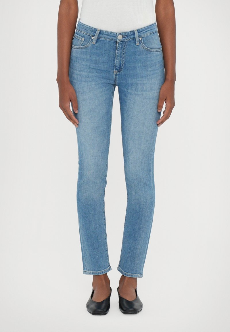 Jean skinny en denim bleu clair avec taille mi-haute, design à cinq poches et décoloration subtile. Des chaussures plates noires complètent la tenue.