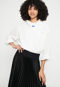 Cămașă albă cu nasturi, cu mâneci suflecate, având un logo și un design zâmbăreț. Asortată cu o fustă plisată neagră, talie elastică și accent cu logo.