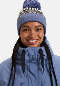 Veste d'hiver bleue avec un col haut, des cordons noirs et un bonnet en tricot présentant un motif géométrique blanc et un pompon bleu.
