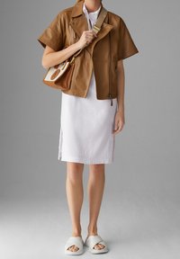 Giacca corta in pelle marrone a maniche corte sopra un vestito bianco. Un paio di sandali in tela leggeri di colore grigio chiaro. L'outfit presenta linee pulite e dettagli minimi.