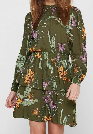 Femme portant une robe verte à motifs floraux, à manches longues, taille élastique, avec des motifs de fleurs jaunes et violettes.