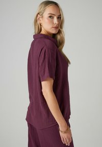 Top corto viola con colletto, realizzato in tessuto texturizzato. Presenta una vestibilità rilassata e sottili righe verticali.