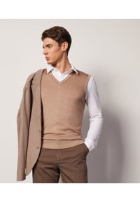 Beżowy sweter bez rękawów z dekoltem w serek noszony na białej koszuli z kołnierzykiem, w zestawieniu z szarym płaszczem i brązowymi spodniami, na neutralnym tle.