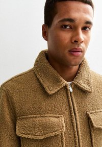 Bruine, gestructureerde jas met een ritssluiting aan de voorkant, twee borstzakken en een kraag. De stof lijkt warm en zacht, geschikt voor koelere weersomstandigheden.