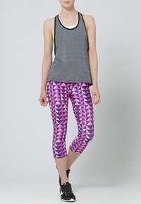 Canottiera grigia con bordi neri sovrapposta a una maglietta gialla chiara, abbinata a leggings rosa con motivo geometrico e scarpe sportive nere.