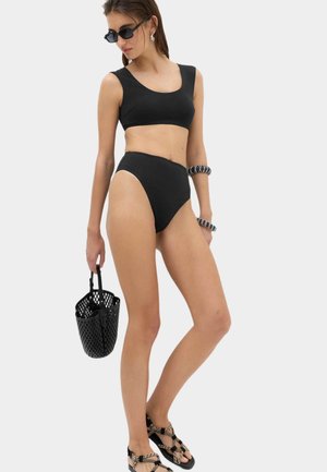 Kobieta w czarnym bikini z wysokim stanem i crop topem, czarnych okularach słonecznych, czarno-białych bransoletkach, trzyma czarną perforowaną torebkę i nosi sandały.
