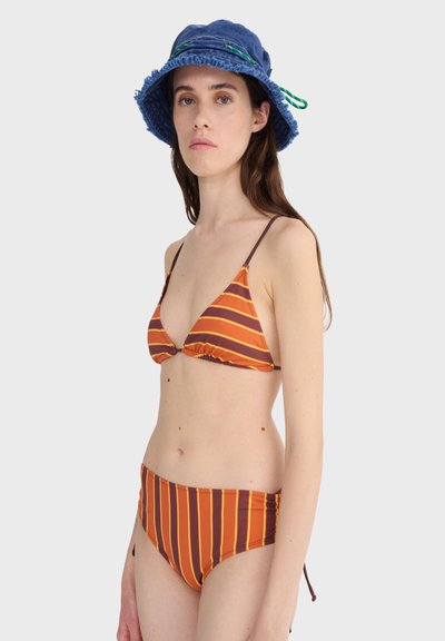 Femme portant un bikini rayé orange et marron et un chapeau seau bleu avec des boucles vertes, debout devant un fond gris clair uni.