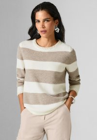 Frau mit langen dunklen Haaren trägt einen beige-weißen gestreiften Pullover, helle beige Hosen, silberne Ohrringe und ein silbernes Armband und posiert mit den Händen in den Taschen.