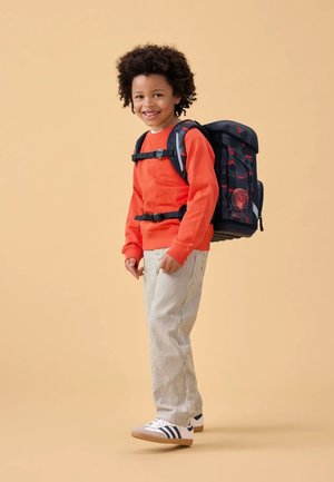Sac à dos à motif noir et rouge avec plusieurs sangles, porté par un enfant en sweat-shirt orange et pantalon rayé. Fond clair.