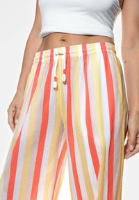 Lekkie, paskowane spodnie w kolorze czerwonym, żółtym i białym. Posiadają elastyczny pas z sznurkiem. Materiał wydaje się być miękki i przewiewny.