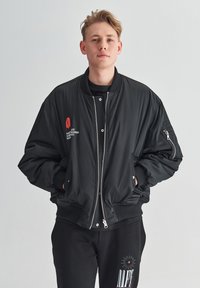 Czarna kurtka bomberka z zamkiem z przodu, ściągaczami przy mankietach i u dołu, z czerwonymi haftowanymi akcentami. Nosi ją osoba w czarnej koszuli i czarnych spodniach.