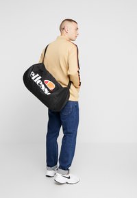 Svart duffelbag med stort hvitt "ellesse"-logo og oransje detaljer, laget av slitesterkt stoff, med buet form og justerbar stropp.