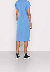 Blaues Midikleid mit kurzen aufgerollten Ärmeln und tailliertem Gürtel. Kombiniert mit schwarzen High-Top-Sneakers mit hellblauen Akzenten.