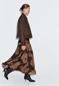 Frau geht und trägt eine braune Wildlederjacke, einen langen, fließenden dunkelbraunen Rock mit beigen Mustern und schwarze Stiefeletten mit Absatz vor einem schlichten Hintergrund.