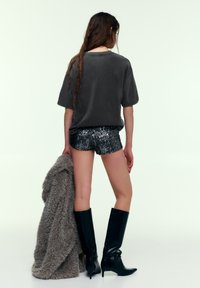 Oversized grå T-shirt, skinnende sorte palietshorts, knælange sorte støvler og en tekstureret grå pelsovertræk hængt over armen.