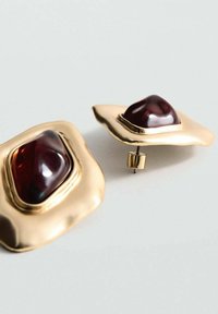 Zlaté náušnice s vlnitým dizajnom, s centrálnym tmavočerveným drahokamom. Hladký povrch s lešteným povrchom, zabezpečujúca zapínacia zátka.