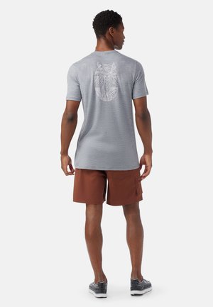 Camicetta grigia a maniche corte con una grafica a contorno sul retro. Indossata con pantaloni corti marroni e sneakers con motivo. Design semplice e vestibilità rilassata.