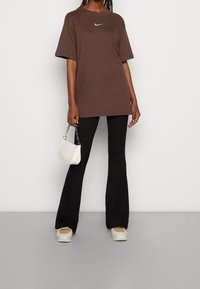 Hnědé oversized tričko Nike s bílým logem, spárované s černými zvonovými kalhotami a bílými teniskami. Malá bílá taška s řetízkovým popruhem.